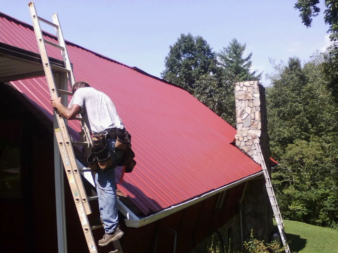 Roofer on a ladder beside a red metal roof and stone chimney — Soffit & Fascia Repair by Roofing Brothers in Westport Point